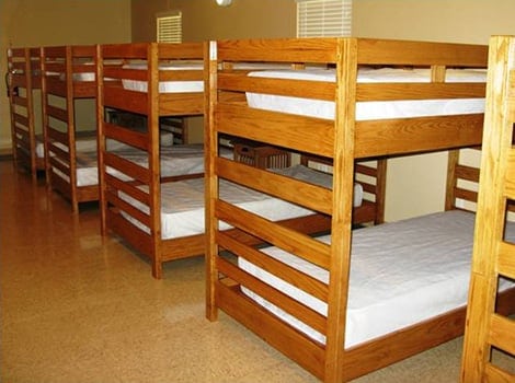 A row of wooden bunk beds with white mattresses in a clean, sparsely furnished room with beige walls and windows.