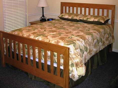A wooden bed with a floral-patterned comforter, two pillows, and a nightstand with a lamp, next to a window with blinds.