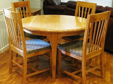 A round wooden dining table with four matching wooden chairs, each with a patterned seat cushion, on a hardwood floor.