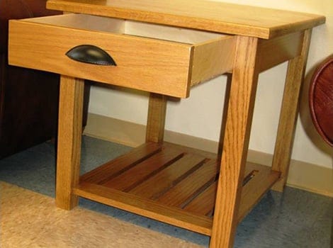 A wooden side table with a single open drawer, a black handle, and a slatted lower shelf standing on a tiled floor.