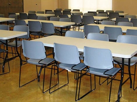 Rows of gray plastic chairs and white rectangular tables are arranged in an empty room with a polished floor.