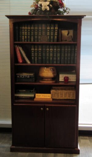A Bookcase with storage doors 36x13x72 displays books, a lamp, a basket, a red apple, and a "Prison Enterprises" sign.