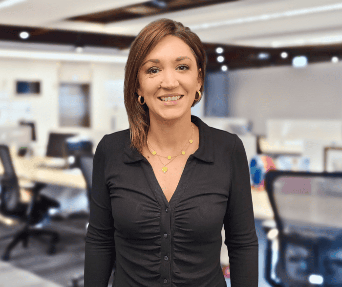 A woman with short brown hair wearing a black top and gold jewelry stands smiling in a modern office setting.