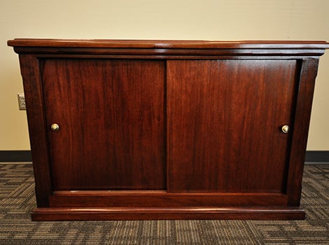 A dark wood cabinet with two sliding doors and round metal handles, placed on a carpeted floor against a beige wall.