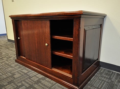 A wooden cabinet with two shelves and a sliding door, placed on a carpeted floor against a light-colored wall.