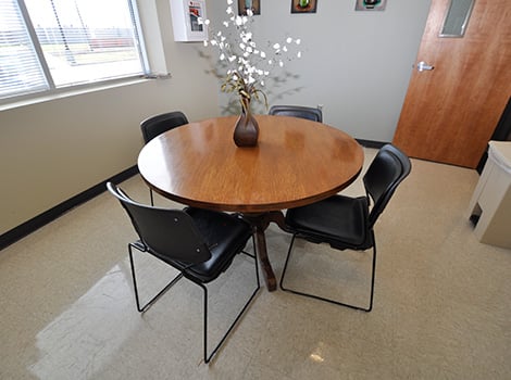 A round wooden table with four black chairs and a brown vase with white artificial flowers, set in a small office room.