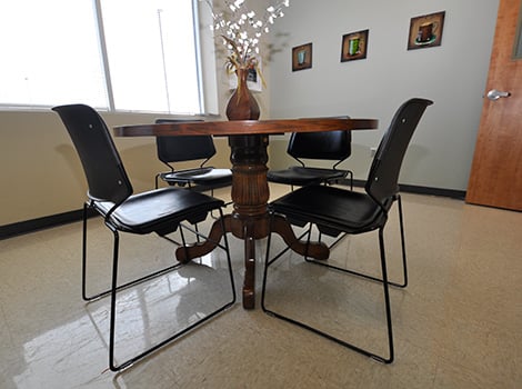 A round wooden table with four black chairs in a room with light walls, a window, and framed pictures on the wall.
