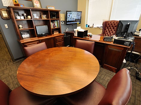A tidy office with a round wooden table, four chairs, a large desk, shelves, and a TV mounted on the wall.