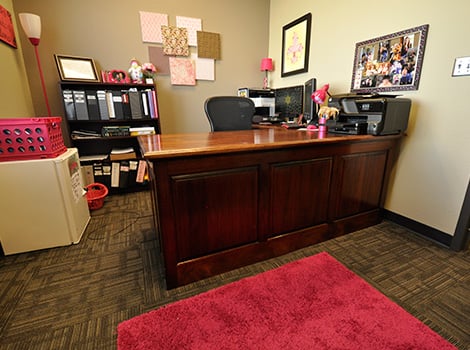 Office with a wooden desk, black office chair, computer, printer, shelves, and pink decor including a rug and various accessories.