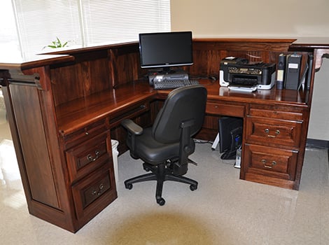 A wooden L-shaped desk with a black office chair, computer monitor, keyboard, printer, and files in an office setting.
