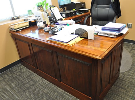 A wooden office desk with papers, folders, a computer, and office supplies; a black chair is behind the desk.