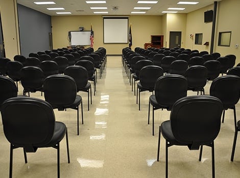 Rows of black chairs are arranged in a conference room facing a projector screen, whiteboard, and two flags at the front.