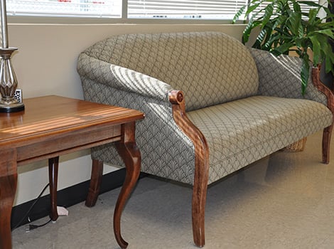 A patterned loveseat with wooden arms sits next to a wooden side table and a potted plant in a room with tile flooring.