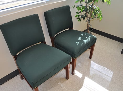 Two green cushioned chairs with wooden legs next to a potted plant in a room with tiled floor and sunlight coming through a window.
