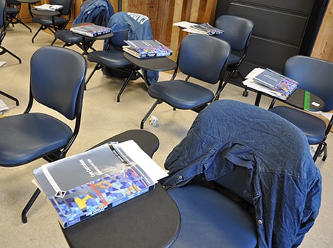 Several empty classroom chairs with attached desks, textbooks, papers, and jackets draped over some chairs are visible in the room.