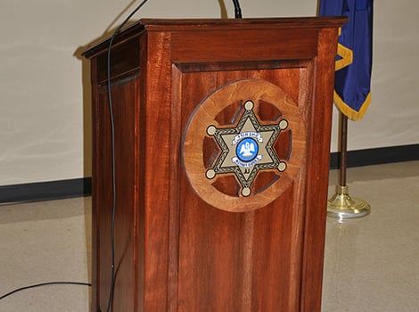 A wooden podium with a sheriff's badge emblem on the front, standing indoors next to a blue and gold flag.