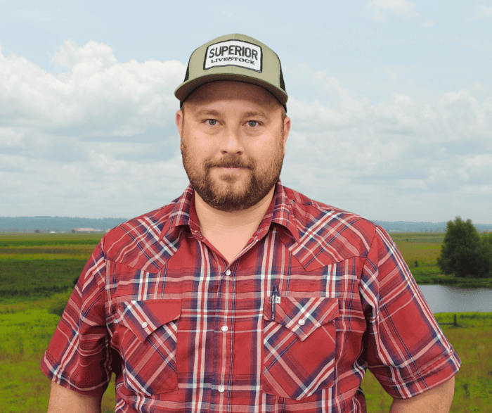 A man in a red plaid shirt and a "Superior Livestock" hat stands outside in front of a grassy field with a pond and cloudy sky.