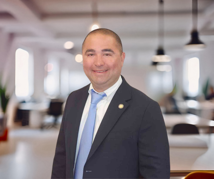 A man in a suit and tie stands smiling in a modern office space with blurred desks and lights in the background.