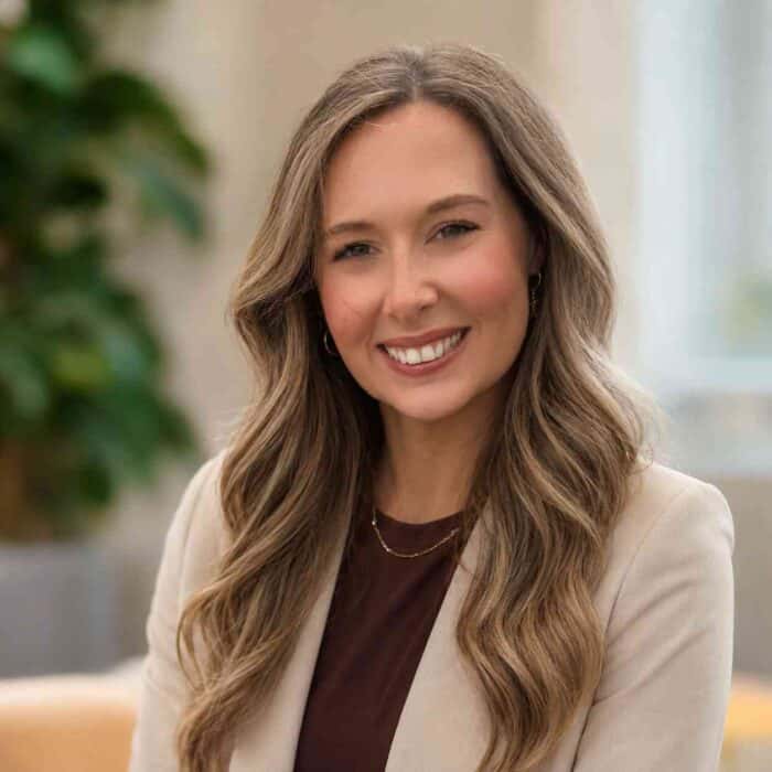 A woman with long wavy hair smiles at the camera, wearing a beige blazer over a brown top, with a blurred background.