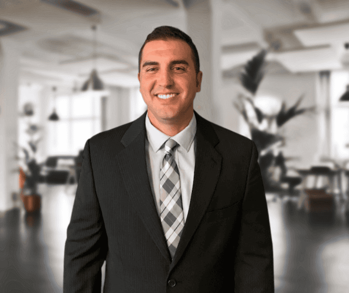 A man in a suit and tie stands smiling in a modern, bright office space with plants and large windows in the background.