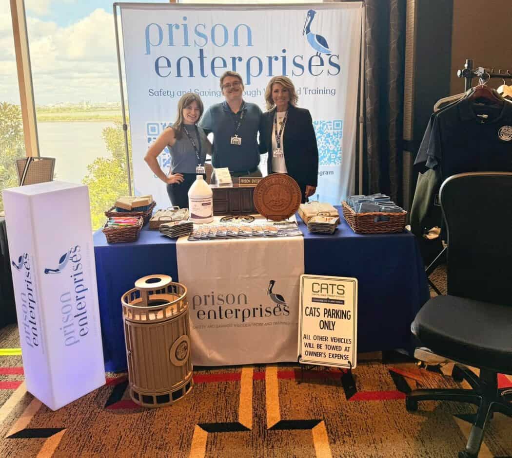 Three people stand behind a Prison Enterprises booth with promotional materials, signage, and a trash bin on display at an indoor event.