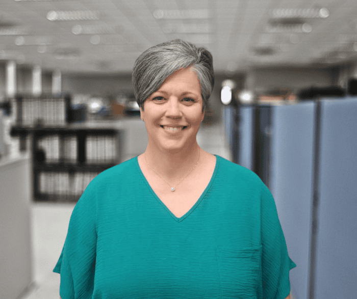 A person with short gray hair and a teal top stands smiling in an office with cubicles and shelves in the background.