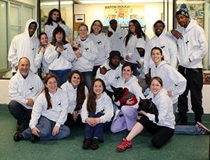 A group of people in white hoodies pose indoors, some holding dogs, with a glass-walled room in the background.