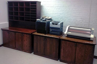 Three wooden cabinets with office equipment on top, including a mail sorter, printers, and a laminating machine, against a white brick wall.