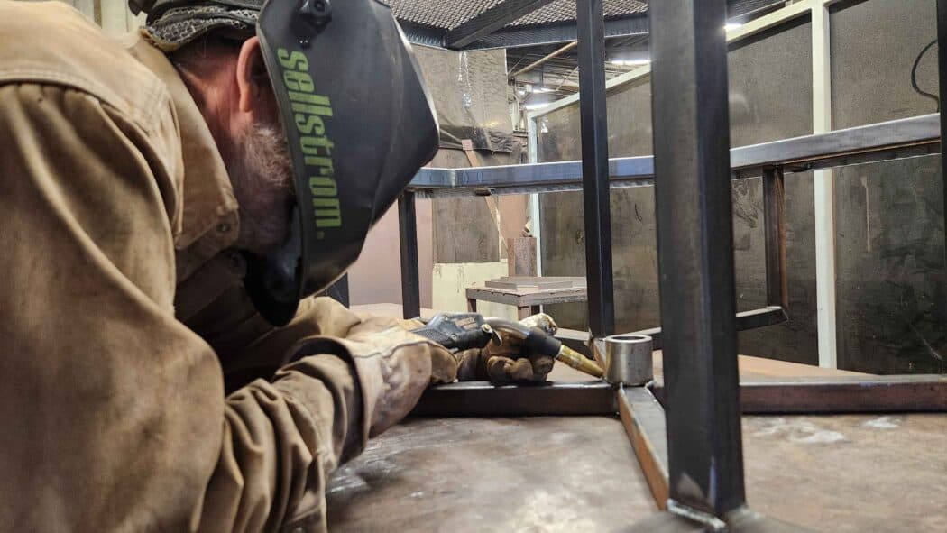 A person in protective gear welds metal frames together in an industrial workshop environment.