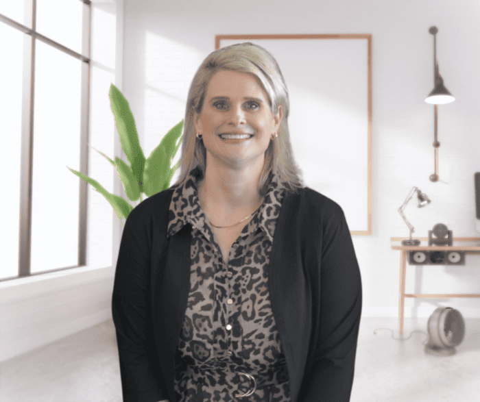 A woman with blonde hair, wearing a black cardigan and patterned blouse, smiles in a modern office with a plant and framed artwork.